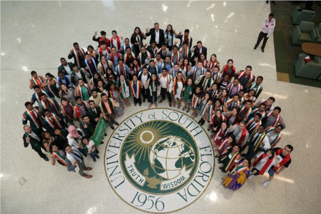 A crowd around the USF seal, looking up to the camera, smiling
